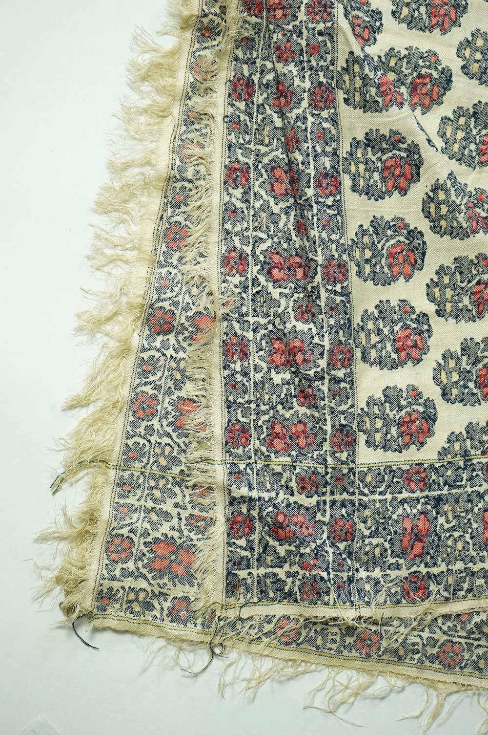 A 19th century silk woven shawl. Woven with a cream silk ground, with indigo, red and yellow, floral stylistic Buta motifs and bordered with three rows of a similar floral vining pattern. The edges fringed. 148cm x 152cm